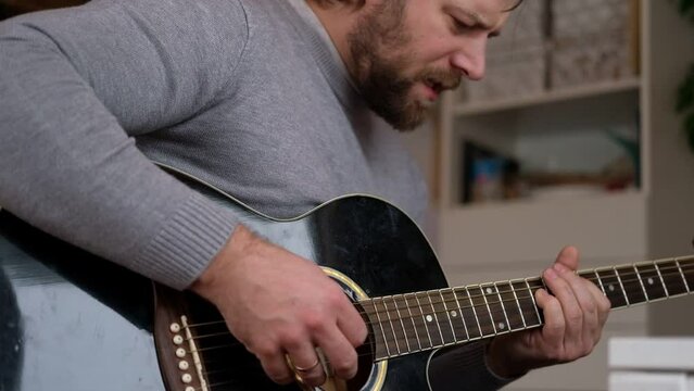 Playing the guitar. Strumming black acoustic guitar. Musician plays music. Man fingers holding mediator. Male hand playing guitar neck in dark room. Unrecognizable person rehearsing, fretboard closeup