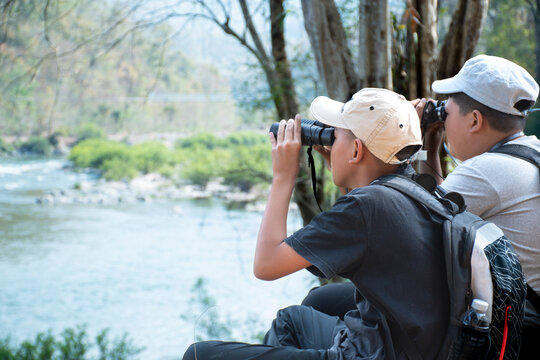 Asian boy sitting by river bank and using binoculars to watch birds on the trees in local national park during summer camp, idea for learning creatures and wildlife animals outside the classroom.