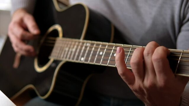 Playing the guitar. Strumming black acoustic guitar. Musician plays music. Man fingers holding mediator. Male hand playing guitar neck in dark room. Unrecognizable person rehearsing, fretboard closeup