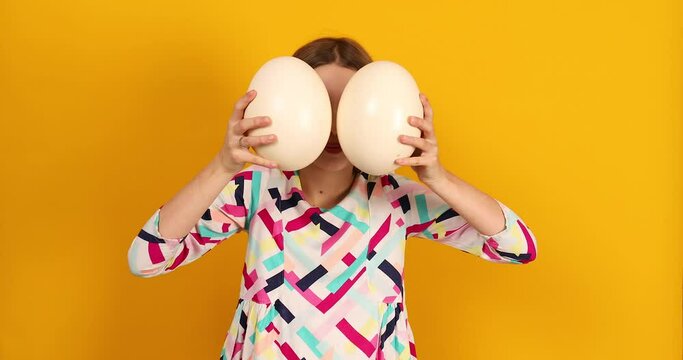 Happy playful teenager girl covering her face Ostrich eggs on a bright yellow studio background, celebrating Easter in style.