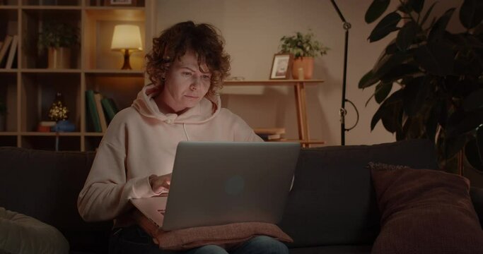 Woman Freelancer Using Laptop While Sitting on Couch at Home. Concentrated Female Person with Curly Hair Working Online Computer and Looking at Device Screen. Concept of Distance Work.