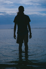 Silhouette of a young long-haired man with his feet in the sea walking towards the shore. Vertical...