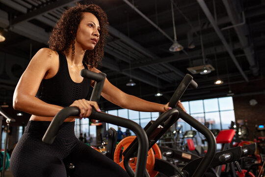 Exercise bike and black woman in the gym for training, cardio training and movement with speed, energy and motivation. Fitness girl, stationary exercise bike and air bike, wellness and action