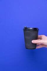 Black paper cup with coffee in a female hand on a blue background, copy space. Front view