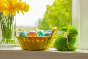 rabbit and basket with colorful eggs on window sill with flowers. Easter symbol, decor, green grass bunny figure