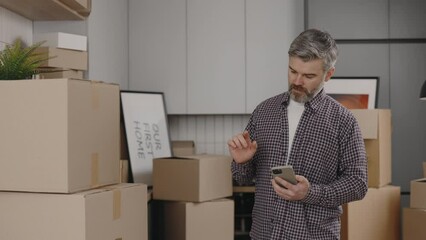 Middle Aged Man Is Using Smartphone While Relocation Standing in New Apartment With Cardboard Boxes on the New Kitchen Background. Male Person Calculating Costs on Mobile.