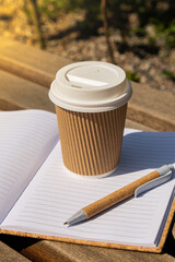 Eco recycling paper cup with coffee or tea on kraft paper with empty paper notebook on wooden bench. Concept of study work outdoors. Take away coffee to go. Copy space for text. Disposable Cardboard