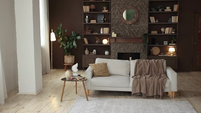 Modern stylish spacious living room with beige sofa, large windows, brown wall, wooden floor, fireplace with mirror near shelving filled with books, decor and potted plants. Cozy chalet interior