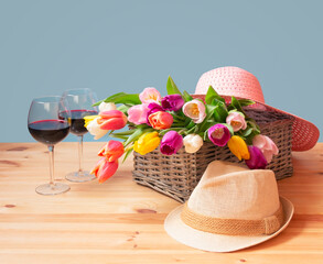 Colorful fresh tulips in wicker basket, hats and glasses of red wine on wooden table.