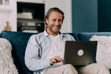 satisfied handsome man 30s with laptop sitting on sofa at home doing online shopping or using...