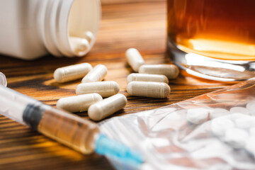 White pills, syringe with a drugs dose, narcotics, glass with alcohol drink on a wooden table, close-up view. Concept of addiction and bad habits