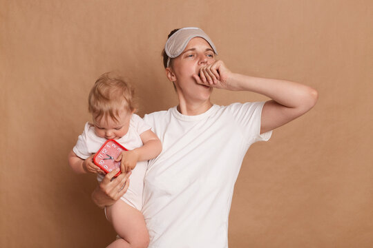 Sleepless woman wearing white t shirt and blindfold, yawning and covering her mouth, holding infant daughter, kid holds red alarm clock, has not enough sleep, posing isolated over brown background.