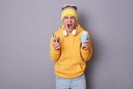 Horizontal shot of angry man wearing beanie hat and yellow hoodie posing against gray wall, holding thermos with hot coffee or tea, using cell phone, yelling with anger and hate.