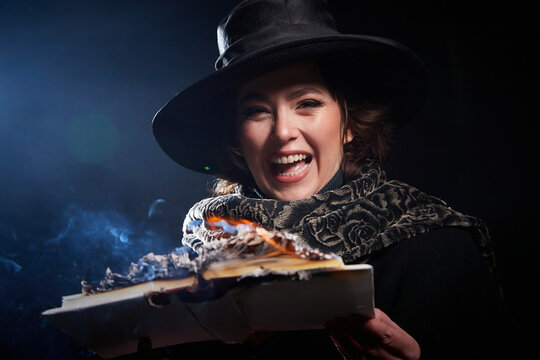 Portrain of witch girl with fier on magic Book. Woman in hat conjuring, making witchcraft over spooky dark black magic background. Model posing in Halloween party. Author writer burns manuscript
