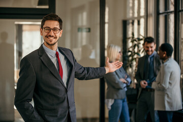 Naklejka na ściany i meble Cropped shot of a businessman standing in the office with his arms pointing to a team, looking confident and smiling at the camera. Group of people talking in the background.