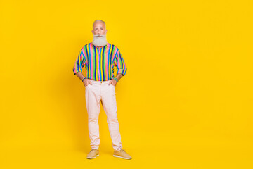 Fototapeta premium Photo of handsome retired man with white beard dressed striped shirt white trousers arms in pockets isolated on yellow color background