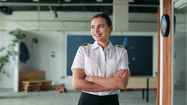 Front view of pretty, female pilot teaching, instruction in classroom. Woman wearing uniform standing with crossed hands, looking at camera, smiling. Concept of enjoying aviation.