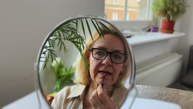 A middle-aged woman with glasses paints her lips with a gloss in front of a mirror. Concept home make-up.