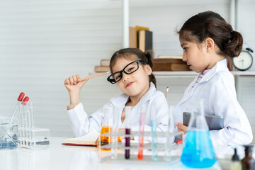 Two little Asian girls in white lab coat help each other for experiment. Little glasses girl holding a pencil to her head and sly smile with new idea while checking recorded information in textbook.