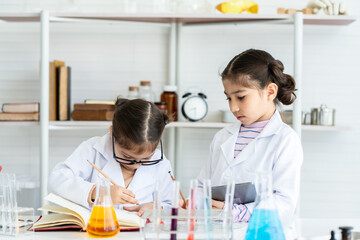 Two little Asian girls in white lab coat help each other for experiment. Little glasses girl writing her new idea by pencil for next experiment in textbook. Her friend looks curious what is her idea?