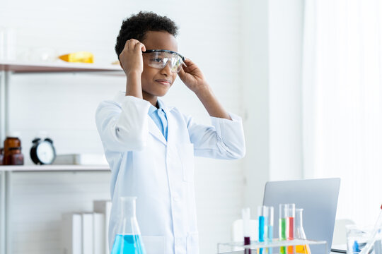 In chemistry classroom with many laboratory tools. A young African boy in white lab coat wearing safety glasses and smile about his new idea for next experiment with many colorful test tubes on table.