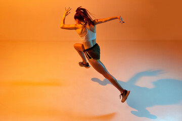 Top side view dynamic image of professional female runner, athlete training over orange studio background in neon light. Concept of sportive lifestyle, health, endurance, action and motion. Ad