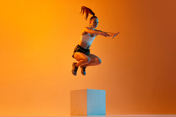 Strong body. Young sportive girl, athlete training, jumping over block against orange studio...