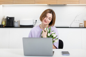 Young happy woman with bouquet of flowers sitting at the table and talking on video call on laptop. Online dating and celebrating holidays far from each other concept