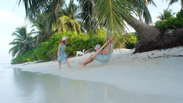 Woman with children having fun on tropical beach. Little girl swinging her mother with baby sister on swings, slow motion