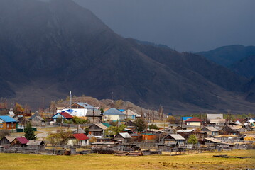 Russia. The South of Western Siberia, the Altai Mountains. The village of Tenga, illuminated by the evening sun, against the background of a snowstorm approaching the rocky mountains.