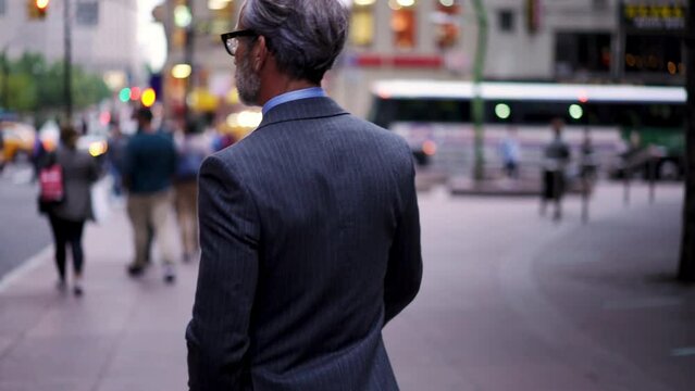 Successful businessman in suit  with cellphone crosses city road t in financial district. Confident entrepreneur in stylish eye glasses walking forward outdoors in downtown. Slow motion 