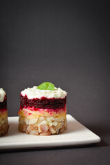 Layered fish salad Herring under fur coat served in culinary rings on dark background. Portion of salad with marinated herring, onion, grated potato, beetroot, mayonnaise on top. Vertical image