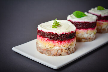 Side view of Layered fish salad Herring under fur coat served in culinary rings on dark background. Portion of salad with marinated herring, onion, grated potato, beetroot, mayonnaise on top