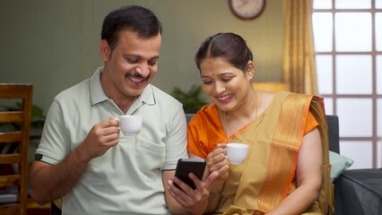 Happy smiling couple drinking tea while watching mobile phone at home - concept of relaxation, refreshment and relationship bonding.
