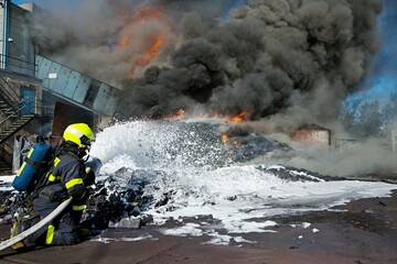 A firefighter uses foam to extinguish a massive plastic waste fire with thick black smoke and flames