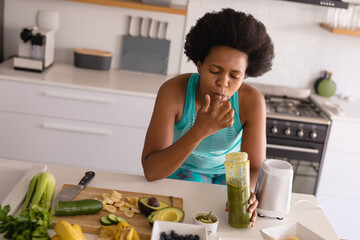 Mid adult african american woman licking finger while drinking healthy juice in kitchen at home
