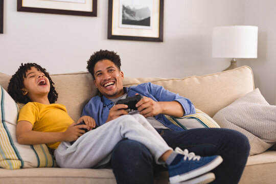 Happy hispanic father and son playing video games together while sitting on sofa in living room
