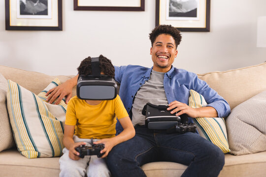 Portrait of hispanic father sitting by son playing video games through vr headset in living room - Powered by Adobe