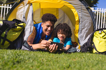 Happy hispanic father and son watching video on smartphone while lying inside tent at backyard