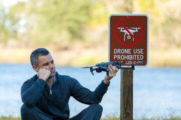 Operator is disappointed because he can not fly his quadcopter in national park no drone area. Man is unable to use his UAV near restriction notice sign - obrazy, fototapety, plakaty