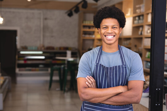 Portrait of happy african american young male owner with arms crossed wearing apron at coffee shop