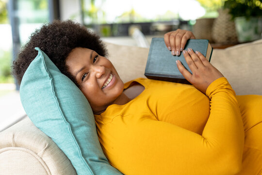 Portrait of smiling african american mid adult woman with book lying on couch at home
