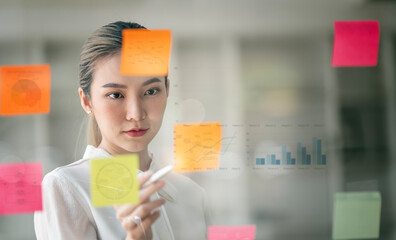 Young smiley attractive businesswoman using sticky notes in glass wall to writing strategy business...