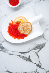 Shovel-shaped plate with red caviar and mini pancakes, above view on a white marble background, vertical shot, copy space