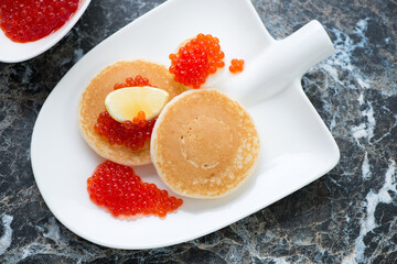Mini pancakes and red caviar served on a shovel-shaped plate, horizontal shot on a black marble background, elevated view