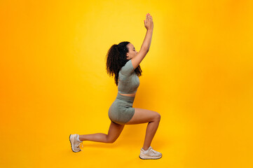 Fototapeta na wymiar Workout concept. Sports lifestyle. Full length photo of a fitness sporty lovely curly haired brazilian or hispanic woman in sportswear, practicing squat exercise and lunges, isolated yellow background