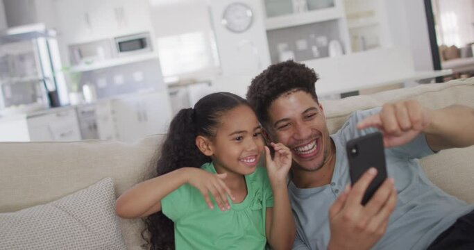 Happy biracial father and daughter sitting on sofa using smartphone