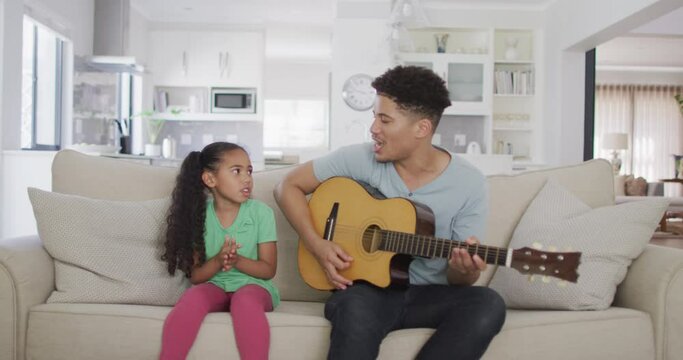 Happy biracial father and daughter sitting on sofa playing guitar