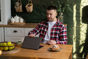 Fototapeta premium A young freelancer in the kitchen sits at a wooden table and closes his laptop after finishing work.