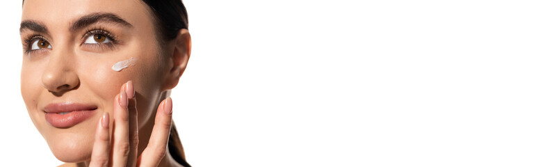 close up view of young woman with face cream on cheek touching face isolated on white, banner.